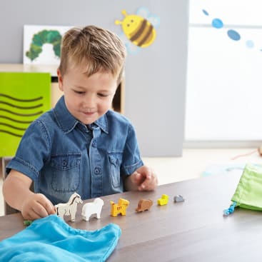 Ein Junge mit einem blauen Hemd sitzt an einem Tisch und ordnet kleine Holztierfiguren in einer Reihe an. Eine grüne Tasche und ein blaues Tuch liegen auf dem Tisch. Im Hintergrund ist ein Klassenzimmer zu sehen.