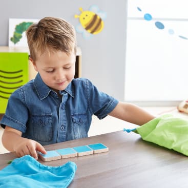 Ein kleines Kind sitzt an einem Tisch, trägt ein blaues Jeanshemd und schaut auf einen Satz blauer Fliesen, während es nach einer grünen Tasche greift. Im Hintergrund sind Dekorationen für das Klassenzimmer, darunter eine Biene und eine Raupe, zu sehen.