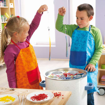 Zwei Kinder mit Schürzen halten selbstgemachte Kerzen hoch, die sie durch Eintauchen von Dochten in einen großen Topf mit Wachs herstellen. Auf dem Tisch daneben liegen verschiedenfarbige Wachsstücke und fertige Kerzen.