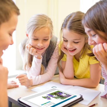 Vier Kinder sitzen um einen Tisch, lächeln und schauen gemeinsam auf einen Tablet-Bildschirm. Auf dem Tisch liegen ein aufgeschlagenes Notizbuch und ein Stift, und das Tablet zeigt eine Webseite mit Bildern und Text an.