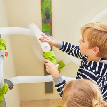Zwei Kinder spielen mit einem an der Wand befestigten Röhrensystem und verbinden flexible Kunststoffröhren mit grünen und orangefarbenen Verbindungsstücken. Der Junge im gestreiften Hemd justiert ein Rohr, während das andere Kind zuschaut.