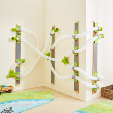 Eine an der Wand montierte Spielstruktur mit durchsichtigen Kunststoffrohren, die durch grüne und graue Armaturen miteinander verbunden sind und ein zusammenhängendes Rutschensystem bilden, ist in der Ecke eines Kinderzimmers mit Spielzeug und Teppichen auf dem Boden installiert.