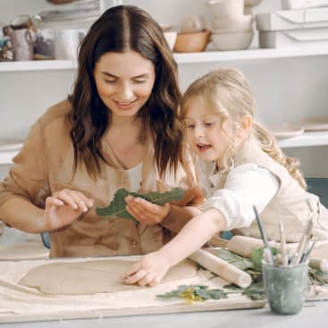 Ein Erwachsener und ein Kind arbeiten an einem Tisch mit Ton und Blättern. Das Kind drückt ein Blatt in den Ton, während der Erwachsene ein anderes Blatt festhält. Im Hintergrund sind Kunstgegenstände und Töpferwaren zu sehen.