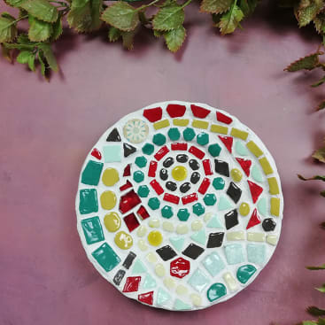 Ein runder Mosaikuntersetzer mit farbenfrohen geometrischen Kacheln in Rot, Grün, Gelb, Schwarz und Weiß sitzt auf einer lilafarbenen Oberfläche, umgeben von grünen Blättern am oberen und rechten Rand.
