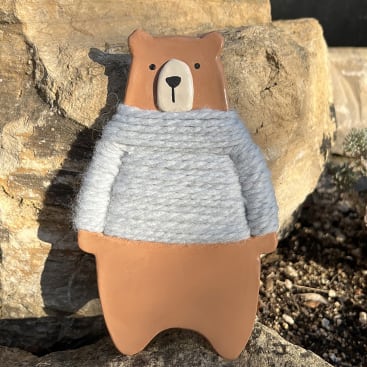 Eine kleine Bärenfigur mit neutralem Gesichtsausdruck, die einen hellgrauen, mit Garn umwickelten Pullover trägt, steht vor einem Hintergrund aus großen Steinen und Kieseln.
