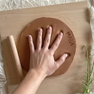 Eine Hand wird auf ein flaches, rundes Stück braunen Tons auf einer hölzernen Unterlage gedrückt, wobei ein kleines Nudelholz in der Nähe liegt. Der Name Maria ist in der Nähe des Randes in den Ton eingraviert. Unten rechts sind weiße Blumen zu sehen.