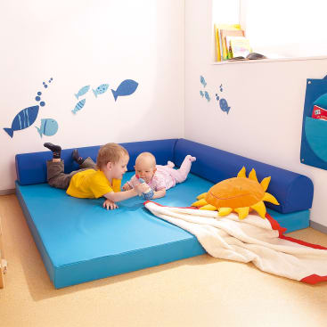 Zwei kleine Kinder liegen auf einer blauen gepolsterten Matte in einer Ecke eines Spielzimmers. In der Nähe befinden sich ein plüschiges Sonnenkissen und eine Decke. Blaue Fischaufkleber zieren die weiße Wand, auf der Fensterbank darüber liegen Bücher.