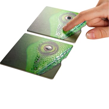Eine Hand hebt eine Karte mit einem grünen, augenähnlichen Muster an, das teilweise zu einer anderen, darunter liegenden Karte auf weißem Hintergrund passt.