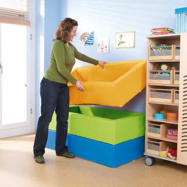 Eine Frau in grünem Hemd und Jeans hebt einen gelben Schaumstoffbehälter aus einem Stapel von drei bunten Behältern in einem hellen Spielzimmer mit Kinderzeichnungen an der Wand und Regalen mit Spielzeug und Büchern.