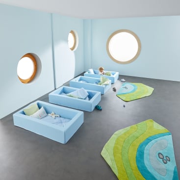 Ein hellblaues Kinderzimmer mit vier kleinen Betten, bunten Kissen und Spielzeug, zwei runden Fenstern und zwei grün gemusterten Teppichen auf einem grauen Boden. In einem der Betten sitzt ein kleines Kind.