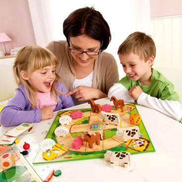 Ein Erwachsener und zwei Kinder sitzen an einem Tisch und spielen ein buntes Brettspiel mit Tierfiguren, darunter Kühe, Schafe, Pferde und Schweine. Die Kinder wirken beschäftigt und glücklich. Würfel und Spielfiguren liegen auf dem Tisch.