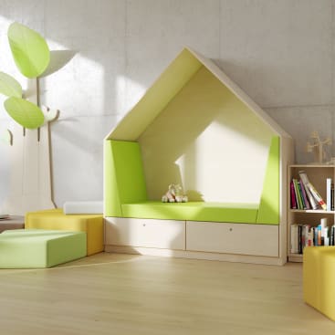 Eine gemütliche Leseecke in Form eines Hauses mit einem grünen Kissen ist an einer Betonwand angebracht. Zwei ausgestopfte Tiere sitzen auf dem Kissen. In der Nähe befinden sich modulare Sitzwürfel, ein Bücherregal mit Büchern und ein baumförmiges Dekorationsstück.