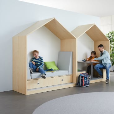 Zwei Kinder und ein Erwachsener sitzen in zwei hölzernen, hausförmigen Nischen; ein Kind sitzt auf einer gepolsterten Bank mit grünen Kissen, während das andere Kind und der Erwachsene an einem Schreibtisch mit Büchern und Heften arbeiten.