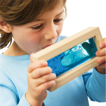 Ein Kind in einem hellblauen Hemd hält ein rechteckiges Objekt mit einem Holzrahmen und einem klaren Fenster, das mit einer blauen Flüssigkeit gefüllt ist, und betrachtet den Inhalt genau.