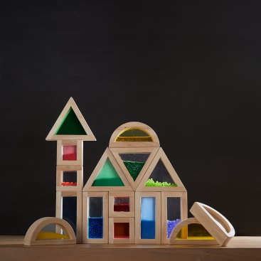 Ein Satz Holzbausteine mit bunten, transparenten Einsätzen ist in Form von Häusern und Bögen vor einem dunklen Hintergrund auf einer Holzfläche angeordnet.