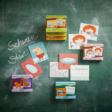 Auf einer grünen Kreidetafel ist eine Sammlung von illustrierten Sprachlernkarten ausgebreitet. Die Karten zeigen Zeichnungen von Kindern, Tieren und Gegenständen. Die deutschen Wörter Schwein und Stier sind mit Kreide geschrieben.
