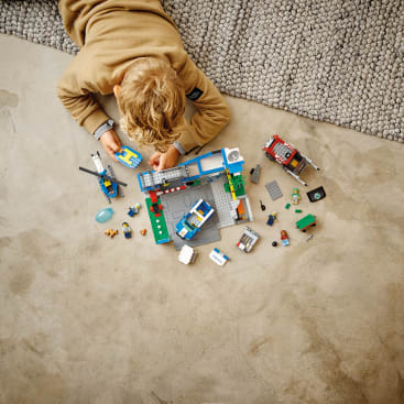 Ein Kind mit blondem Haar und hellbraunem Pullover liegt auf einem Betonboden neben einem grauen Teppich und spielt mit verschiedenen bunten LEGO Steinen und Fahrzeugen, die vor ihm ausgebreitet sind.
