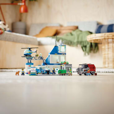 Auf dem Boden steht eine LEGO Polizeistation mit einem Hubschrauber, mehreren Polizeifahrzeugen, einem Polizeihund, Minifiguren und einem kleinen Gefängnis vor einem verschwommenen Wohnzimmerhintergrund.