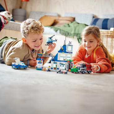 Zwei Kinder liegen auf dem Boden und spielen mit einer LEGO Polizeistation, die kleine Fahrzeuge und Figuren enthält. Das Zimmer hat eine neutrale Einrichtung mit Kissen und einem Weidenkorb im Hintergrund.