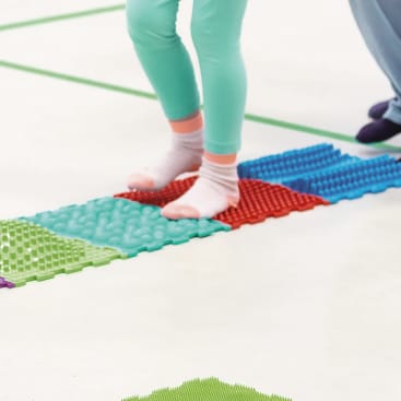 Ein Kind, das mintgrüne Leggings und weiße Socken trägt, läuft auf strukturierten, farbenfrohen Sinnesmatten in einem Raum. Die Beine einer anderen Person sind rechts teilweise sichtbar. Der Fokus liegt auf den Füßen und den Matten.