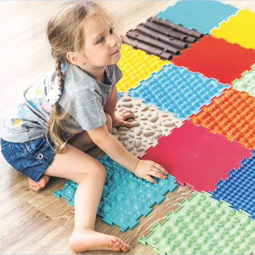 Ein junges Mädchen sitzt barfuß auf einem Holzboden und berührt strukturierte, bunte, ineinandergreifende Sinnesmatten. Die Matten haben verschiedene erhabene Muster und Farben. Das Mädchen trägt ein graues T-Shirt und Jeansshorts.