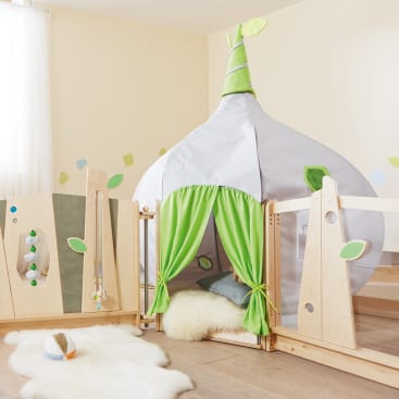 Ein Spielbereich für Kinder mit einem Zelt in Form einer Zwiebel mit grünen Vorhängen, umgeben von hellen Holzpaneelen, einem weißen Fellteppich, Kissen und einem Stofftier auf einem Holzboden.