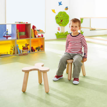 Ein kleines Kind in einem gestreiften Hemd und einer grauen Hose sitzt auf einem kleinen Holzhocker in einem hellen Klassenzimmer mit Regalen voller Spielzeug, Büchern und buntem Wandschmuck.