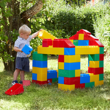 Ein kleines Kind steht im Freien auf einer Wiese und spielt mit großen, bunten, ineinander greifenden Plastikblöcken, um ein hausähnliches Gebäude unter einem Baum zu bauen.