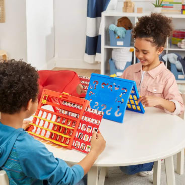 Zwei Kinder sitzen an einem runden Tisch und spielen das Brettspiel Guess Who, wobei das eine ein rotes und das andere ein blaues Brett benutzt. Im Hintergrund sind Spielzeuge und Regale zu sehen.