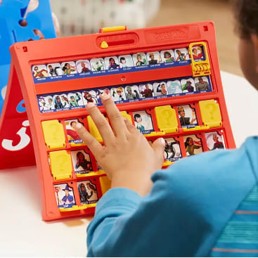 Die Hand einer Person berührt ein rotes und gelbes Spielbrett auf einem Tisch, auf dem "Guess Who? Auf dem Brett sind verschiedene Charakterkarten mit illustrierten Gesichtern und Namen abgebildet. Ein weiteres blaues Spielbrett ist teilweise im Hintergrund zu sehen.