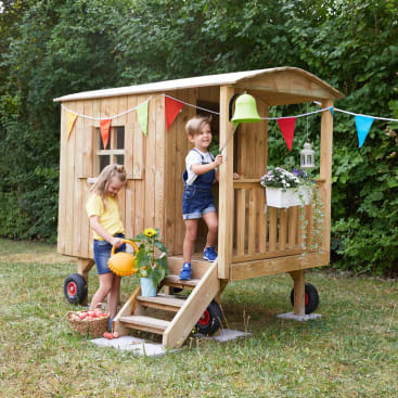 Zwei Kinder spielen draußen mit einem Holzspielhaus auf Rädern, das mit bunten Wimpeln geschmückt ist. Ein Kind läutet eine grüne Glocke, während das andere einen Kürbis hält und neben einem Korb mit Äpfeln steht.