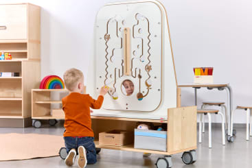 Ein kleines Kind kniet auf dem Boden und spielt mit einem vertikalen, interaktiven Labyrinthbrett in einem Klassenzimmer. Das Gesicht eines anderen Kindes ist durch ein rundes Fenster in der Tafel zu sehen. Im Hintergrund befinden sich Regale, Tische und Stühle.