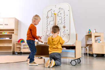 Zwei kleine Kinder spielen in einem Klassenzimmer mit einer großen, vertikalen Holztafel. Im Hintergrund sind Regale mit Spielzeug und Unterrichtsmaterial zu sehen.