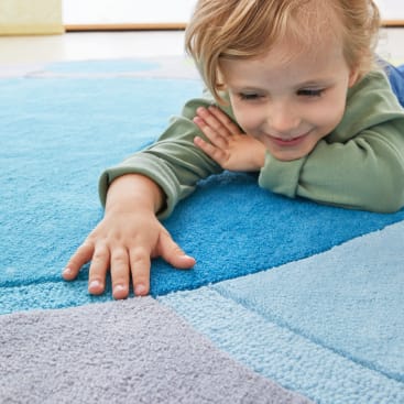 Ein kleines Kind mit hellem Haar und einem grünen langärmeligen Hemd liegt auf einem bunten blau-grauen Teppich, lächelt und berührt den Teppich mit einer Hand.