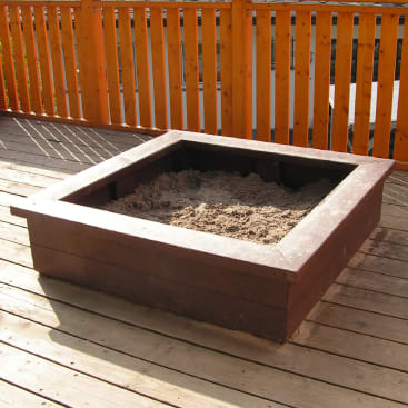 Ein mit Sand gefüllter Holzsandkasten steht auf einem Holzdeck neben einem Holzzaun mit vertikalen Latten.