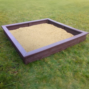Ein quadratischer Sandkasten, gefüllt mit hellem Sand, steht auf grünem Gras. Der Sandkasten hat einen dunkelbraunen Holzrahmen und befindet sich im Freien auf einer offenen Grasfläche.