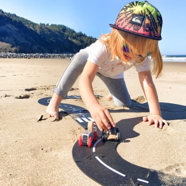 Ein Kind mit bunter Mütze und Sonnenbrille spielt mit Spielzeugautos auf einer Bahn am Sandstrand, im Hintergrund sind Bäume und das Meer zu sehen.