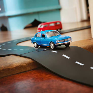 Ein blaues Spielzeugauto und ein roter Spielzeug-Van fahren auf einer kurvenreichen schwarzen Spielzeugstraße mit weißen Markierungen, die auf einer Holzfläche in einem Innenraum steht.