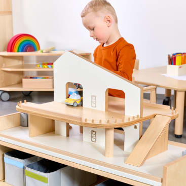 Ein kleines Kind in einem orangefarbenen langärmeligen Hemd spielt mit einem Holzspielzeug-Garagenset, das eine Rampe und ein kleines Spielzeugauto enthält. Im Hintergrund sind Regale mit buntem Spielzeug und Kunstzubehör zu sehen.
