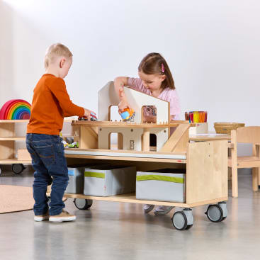 Ein Junge und ein Mädchen spielen mit Spielzeug auf einem hölzernen Aktivitätswagen mit Ablagefächern und Rädern in einem Klassenzimmer. Im Hintergrund sind Kunstgegenstände und ein Regenbogenstapler zu sehen.