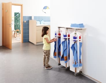 Ein kleines Kind steht vor einem Regal mit blauen Handtüchern und Flaschen und greift nach einem beschrifteten Haken. Die Szene spielt in einem hellen, aufgeräumten Klassenzimmer oder in einer Kindertagesstätte mit einer offenen Tür zu einem anderen Raum.