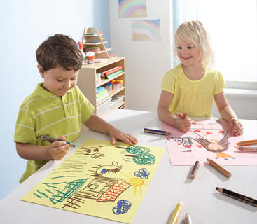 Zwei kleine Kinder sitzen an einem Tisch und zeichnen mit Markern. Der Junge malt auf gelbem Papier mit Naturmotiven, während das Mädchen lächelt und auf rosa Papier malt. Im Hintergrund sind Malutensilien und bunte Kunstwerke zu sehen.
