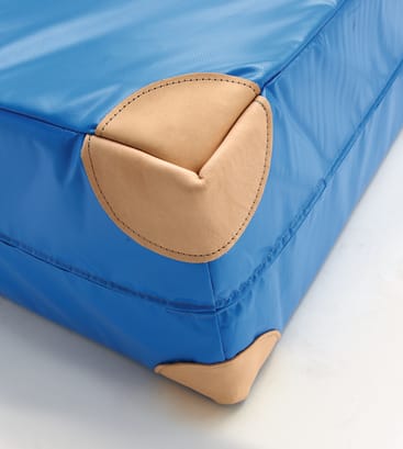 Nahaufnahme einer blauen, gepolsterten Matte mit hellbraunen, verstärkten Ecken aus einem anderen Material, die Details der Nähte zeigt. Die Matte liegt auf einer hellen Unterlage.