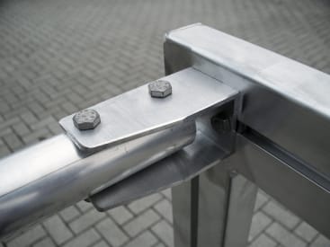 Nahaufnahme einer Metallverbindung, die mit zwei Schrauben an einer Metallstruktur befestigt ist, mit einer gepflasterten Fläche im Hintergrund.