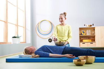 Ein Mädchen liegt mit geschlossenen Augen auf einer Yogamatte, während ein anderes Mädchen eine Klangschale über ihren Bauch hält und anschlägt. Mehrere Klangschalen stehen in der Nähe in einem hellen Raum mit Hula-Hoop-Reifen und Regalen im Hintergrund.