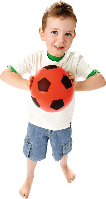 Ein Junge steht barfuß, trägt ein weißes T-Shirt mit grünem Rand und blaue Shorts. Er hält einen rot-schwarzen Fußball und lächelt in die Kamera vor einem schlichten weißen Hintergrund.