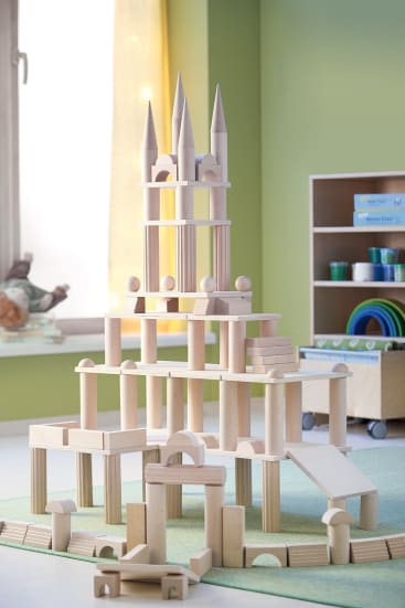 Ein großes, kompliziertes Gebilde aus Holzbausteinen steht auf einer grünen Matte in einem Spielzimmer mit Regalen, Spielzeugaufbewahrung und einem Fenster mit gelben Vorhängen im Hintergrund.