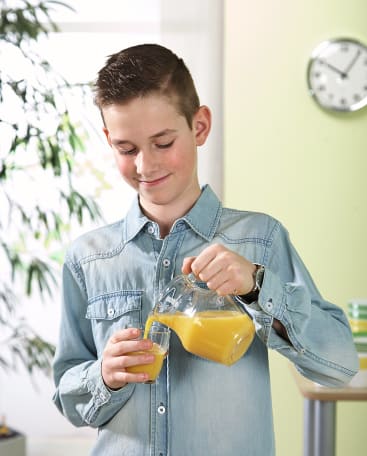 Ein Junge in einem hellen Jeanshemd gießt Orangensaft aus einem Glaskrug in ein kleines Glas. Im Hintergrund sind eine Pflanze, eine Uhr und einige gestapelte Teller zu sehen.