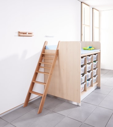 Eine Wickelkommode aus hellem Holz mit einer Leiter, einer Wickelauflage, Babyzubehör oben und 12 beschrifteten Aufbewahrungsbehältern unten, auf einem gefliesten Boden vor einer weißen Wand.