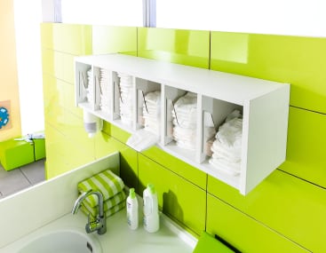 Ein weißer, an der Wand befestigter Organizer über einem Waschbecken, der Stapel von Windeln in mehreren Fächern enthält, mit Babyflaschen, Seife und Handtüchern auf dem Tresen. Die umliegenden Fliesen sind hellgrün.